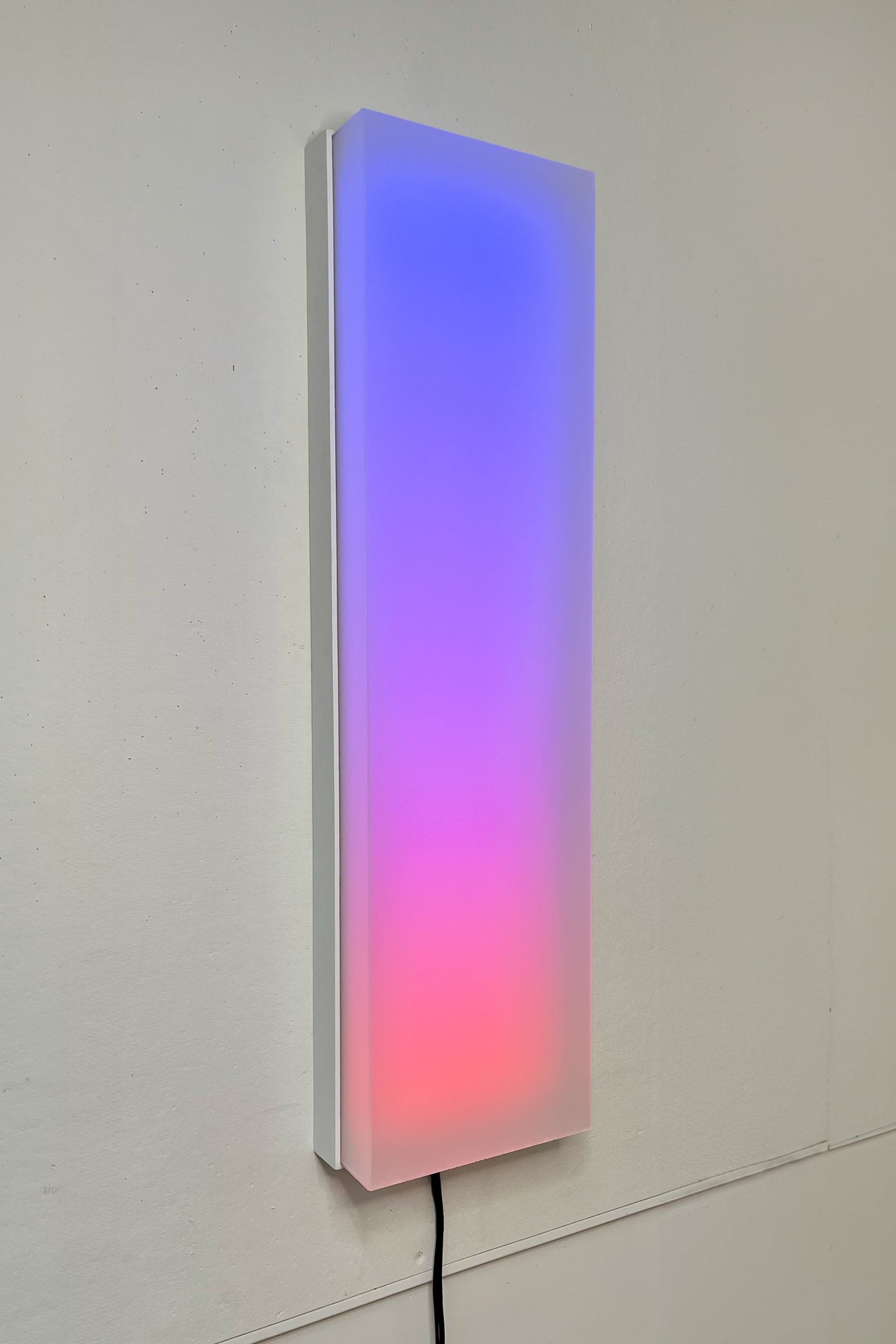 Historical Circadia prototype on a pale wall, with a blue-to-pink luminous face, exposed side and a visible black power cable.